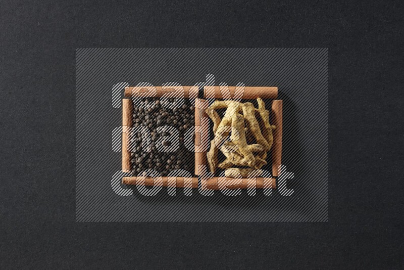 مربعان من أعواد القرفة ممتلئان بحبوب الفلفل الأسود وأصابع الكركم علي خلفية سوداء