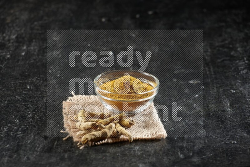 A glass bowl full of turmeric powder with dried turmeric fingers on a burlap fabric on textured black flooring