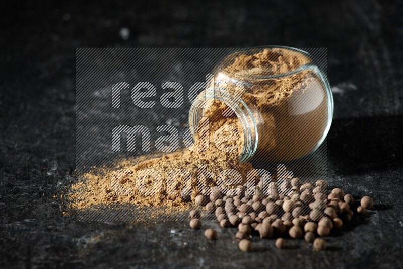 A flipped glass spice jar full of allspice powder and powder spilled out of it with whole balls on a textured black flooring