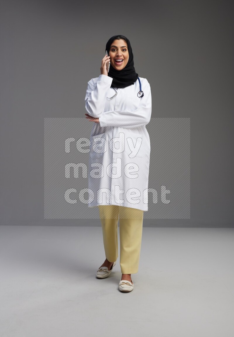 Saudi woman wearing lab coat with stethoscope standing talking on phone on Gray background