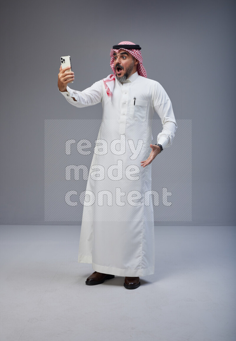 Saudi man Wearing Thob and red Shomag standing taking selfie on Gray background