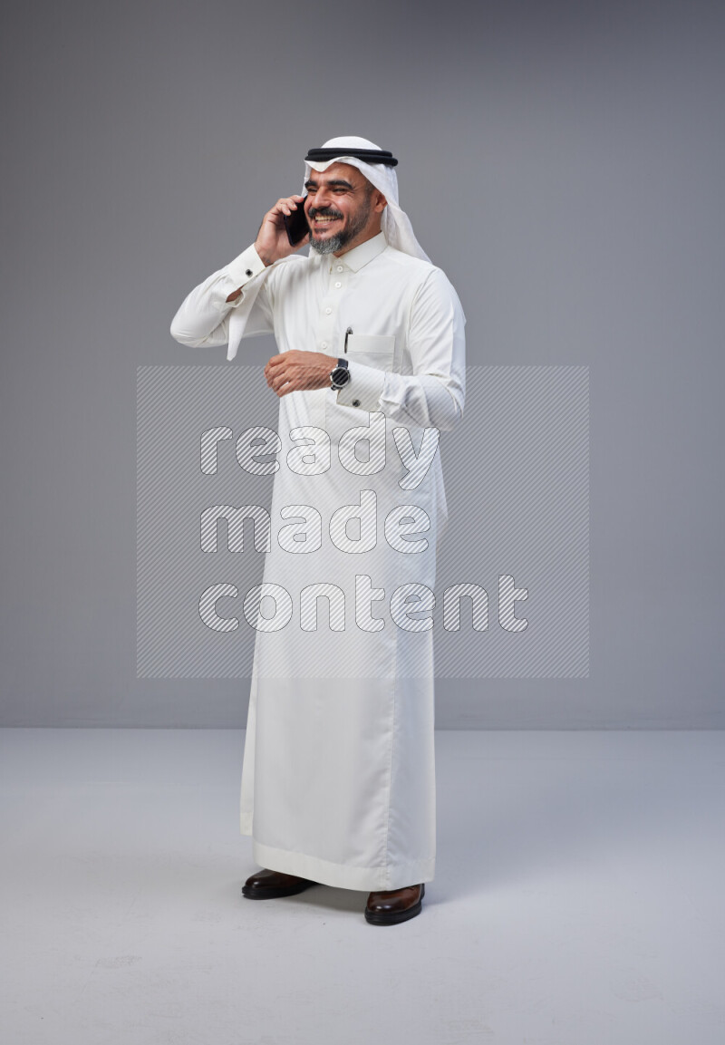 Saudi man Wearing Thob and white Shomag standing talking on phone on Gray background