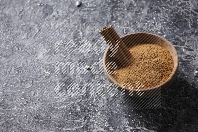 Wooden bowl full of cinnamon powder and a cinnamon stick on a textured black background