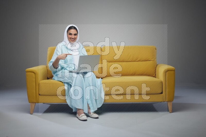 A Saudi woman sitting on a yellow Sofa With her Laptop Pointing wearing Blue Abaya
