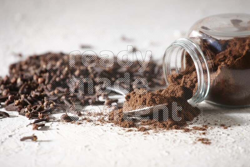 A flipped glass spice jar and a metal spoon full of cloves powder and powder came out of the jar with cloves spread on textured white flooring