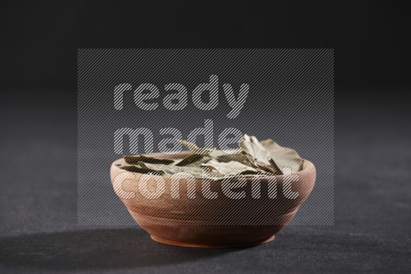 A wooden bowl filled with dried bay leaves on black flooring