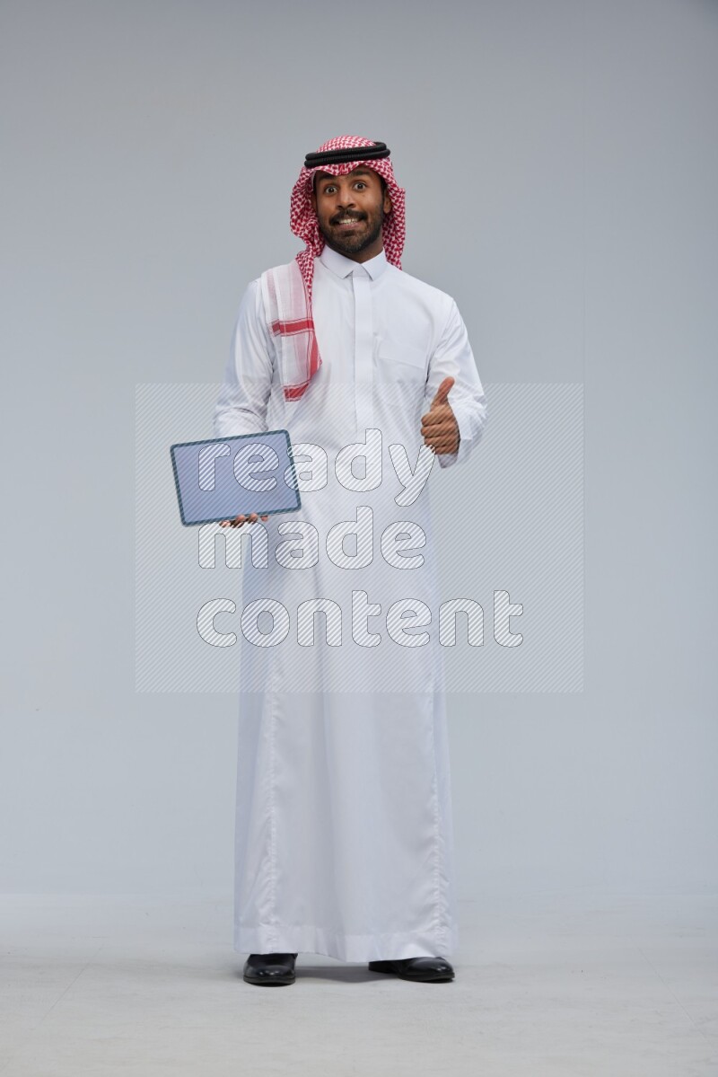 Saudi man Wearing Thob and shomag standing showing tablet to camera on Gray background