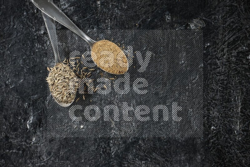 2 metal spoons full of cumin seeds and cumin powder on a textured black flooring