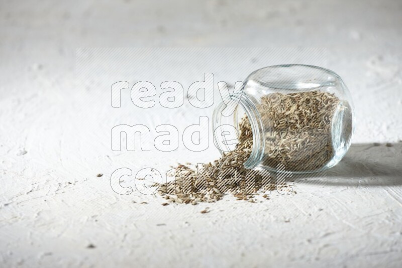 A flipped glass spice jar full of cumin seeds and the seeds spilled out on textured white flooring