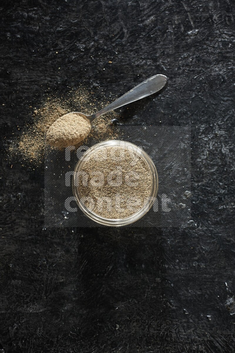 A glass jar and metal spoon full of cardamom powder on textured black flooring