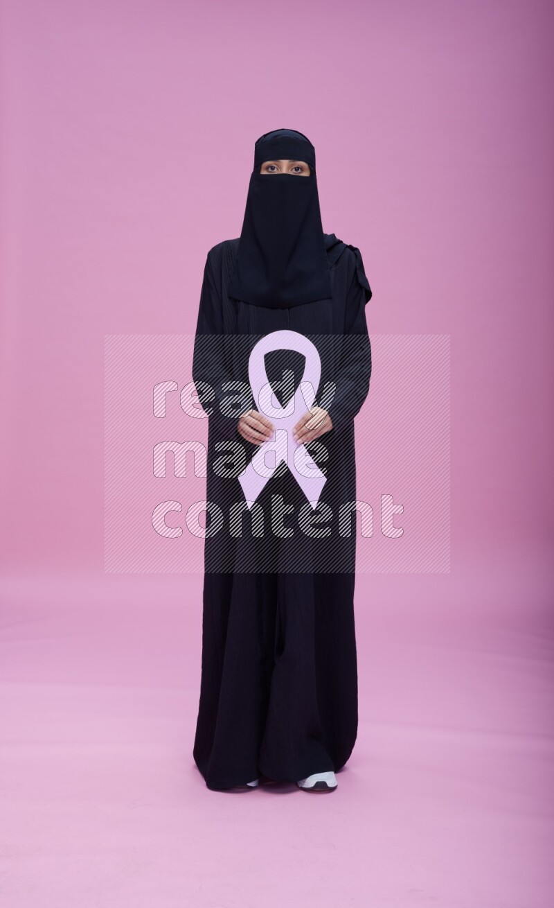 Saudi woman wearing abaya and niqab standing holding awareness ribbon on pink background
