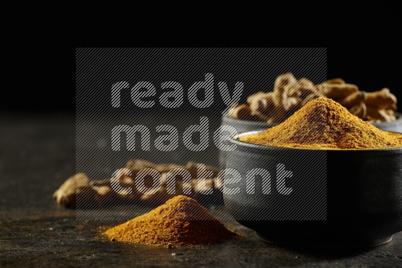 2 pottery black bowls, one full of turmeric powder and the other full of dried turmeric whole fingers on a textured black flooring