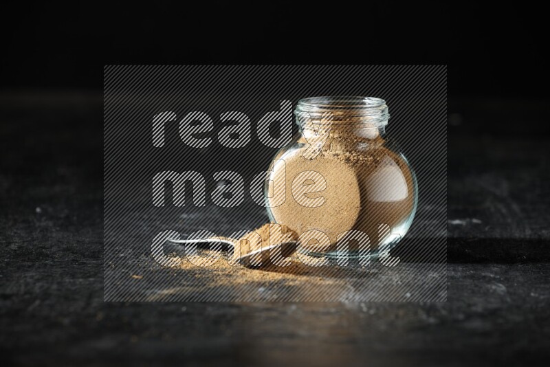 A glass spice jar and metal spoon full of allspice powder on a textured black flooring