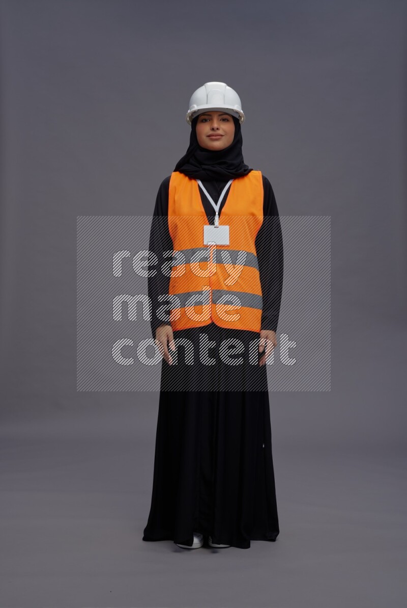 Saudi woman wearing Abaya with engineer vest with neck strap employee badge standing interacting with the camera on gray background