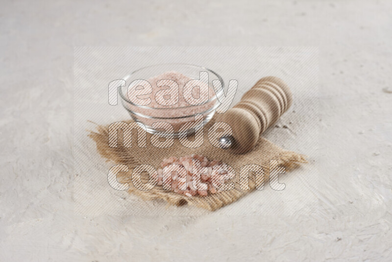 A glass bowl full of pink himalayan salt with a wooden grinder on a burlap fabric all on white background