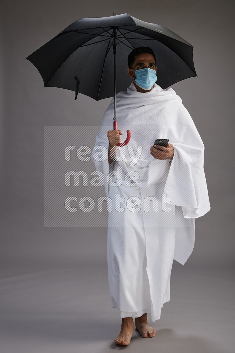 A man wearing Ehram and mask Standing holding umbrella on gray background on gray background