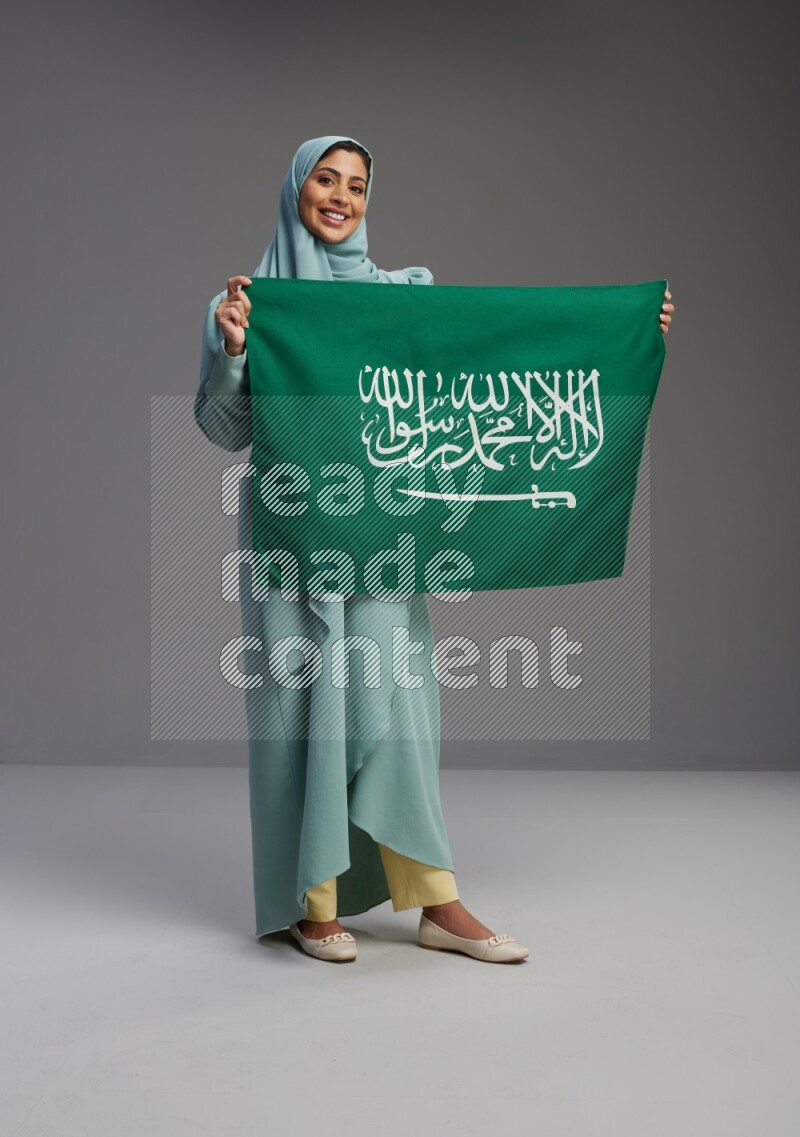 Saudi Woman wearing Abaya standing holding Saudi flag on Gray background