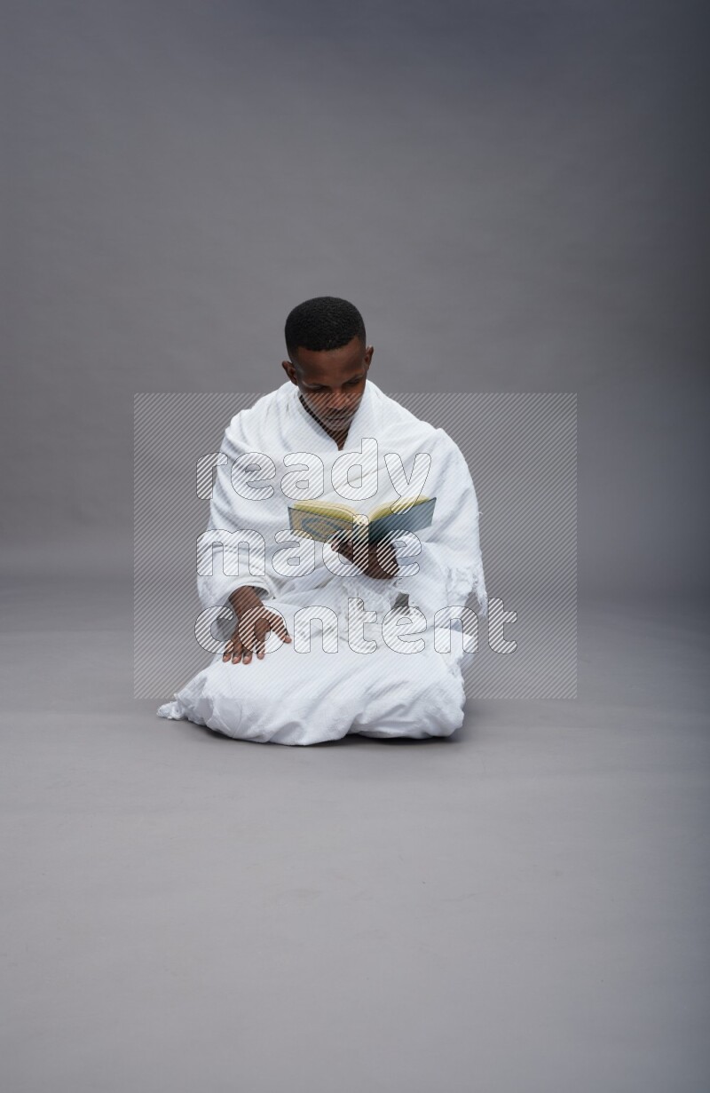 A man wearing Ehram sitting on floor reading quran on gray background