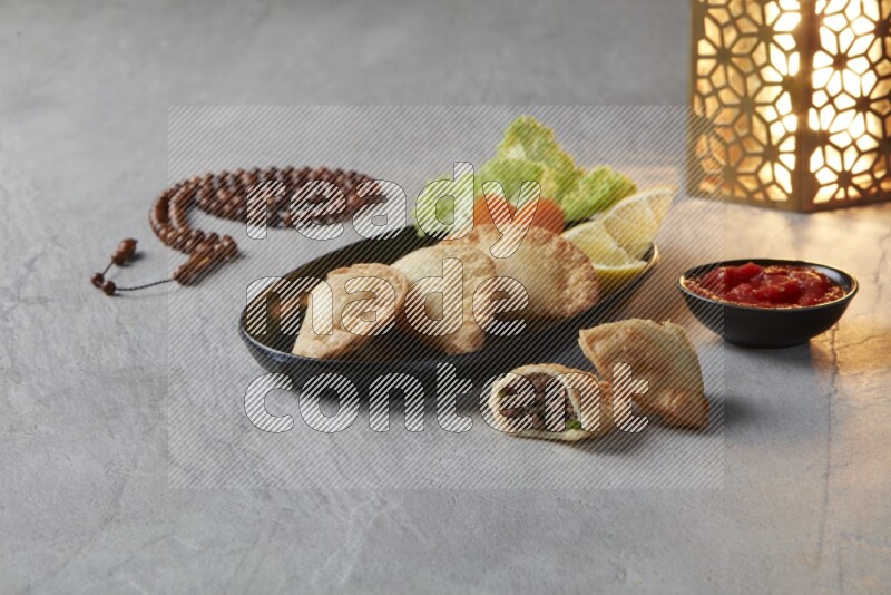 Three fried sambosas in an oval shaped black plate and a red sauce in a black round ramekin with a brown misbaha and a golden lantern on the side on a gray background