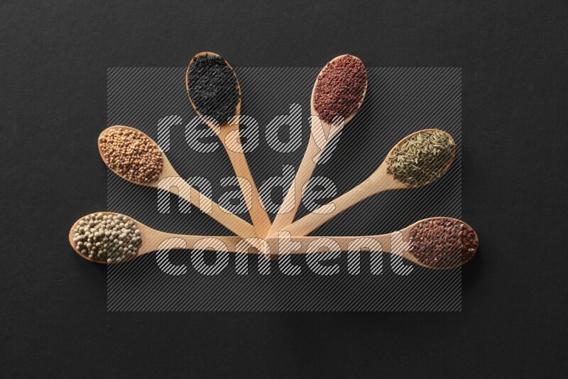 wooden spoons filled with white peppers, mustard seeds, black seeds, garden cress, cumin and flax on black flooring
