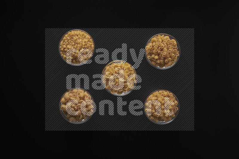 Different pasta types in 5 glass bowls on black background