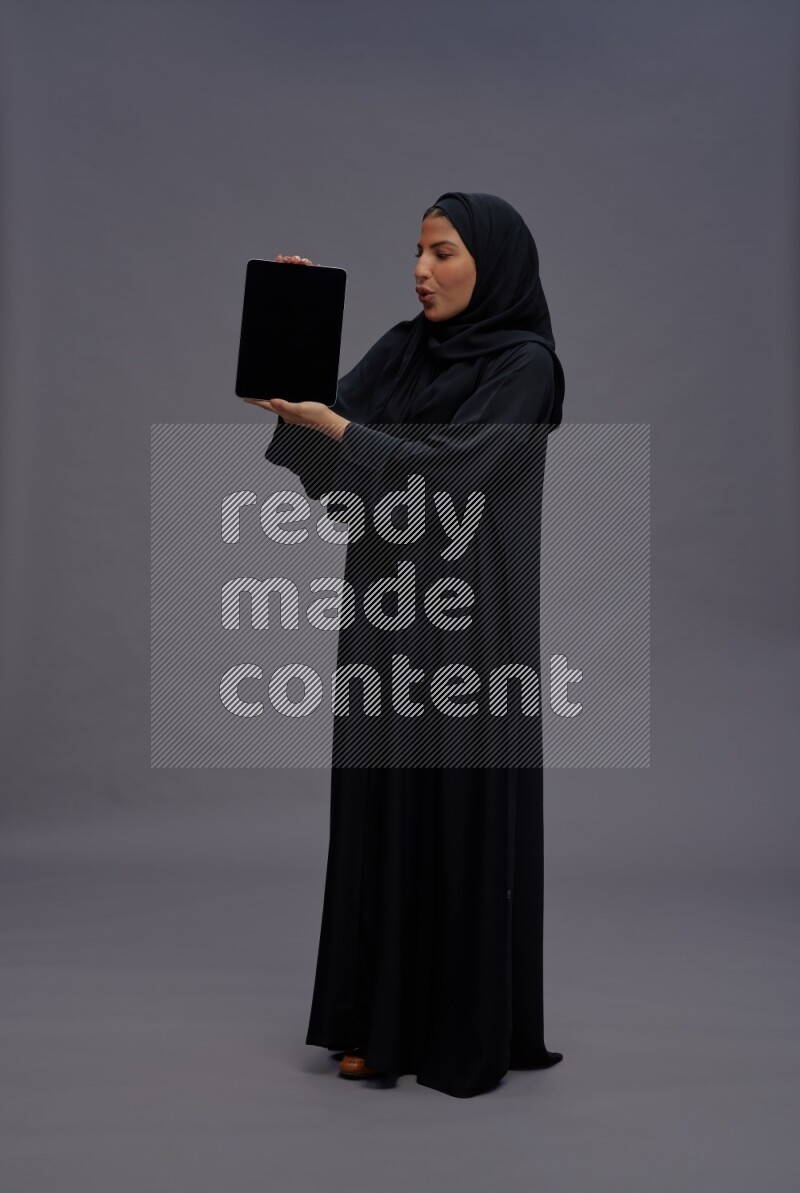 Saudi woman wearing Abaya standing showing tablet to camera on gray background