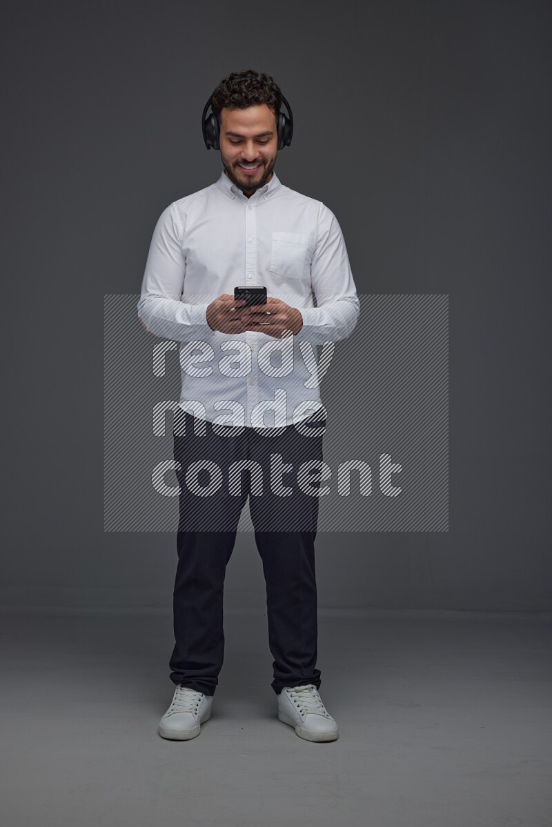 A man wearing smart casual and using his phone and headphone eye level on a gray background