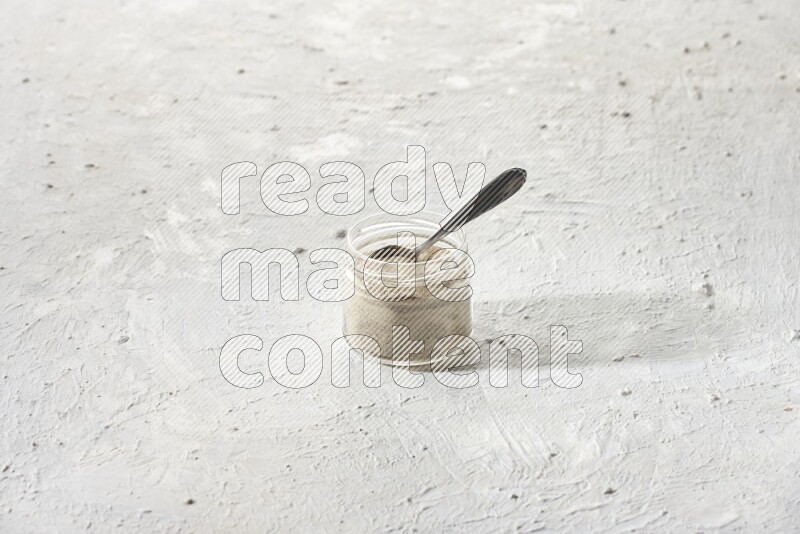 A glass jar and a metal spoon full of white pepper powder on textured white flooring