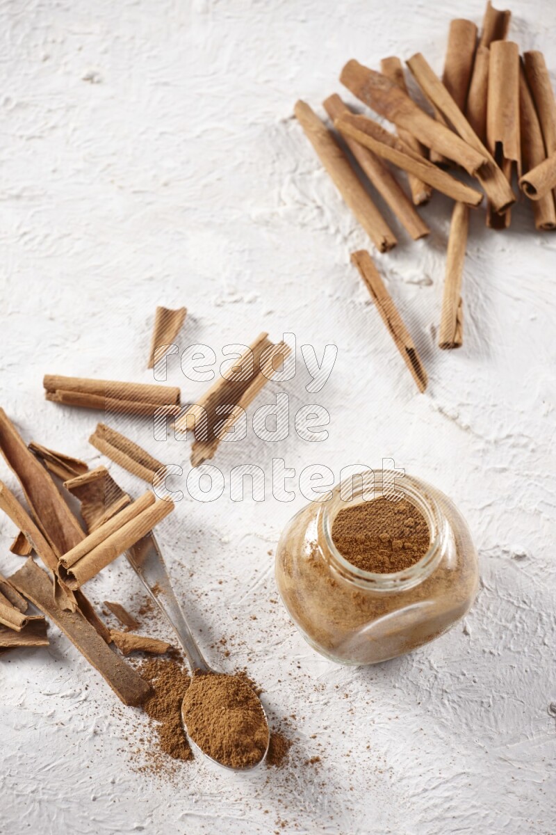 Herbal glass jar full cinnamon powder and a metal spoon surrounded by cinnamon sticks on a white background