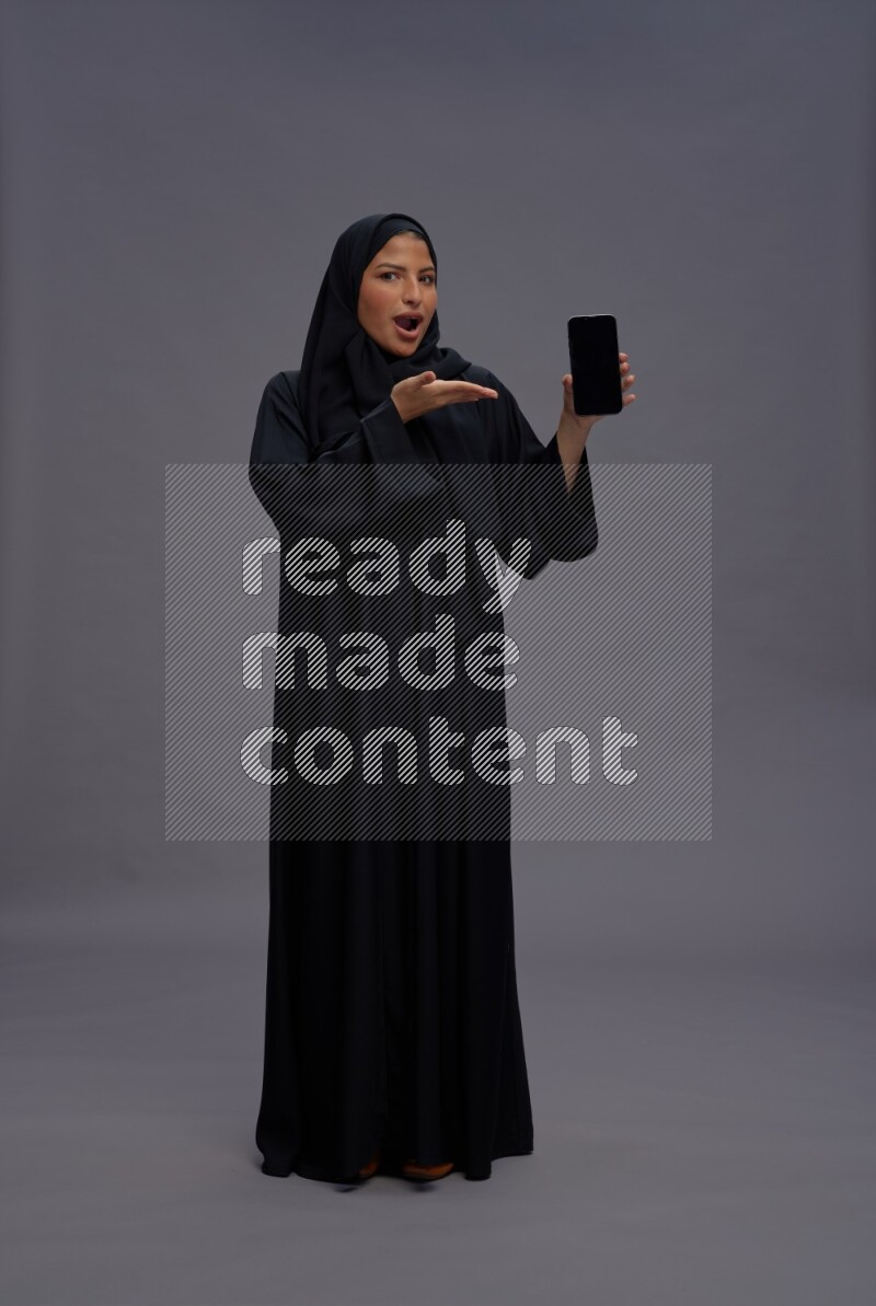 Saudi woman wearing Abaya standing showing phone to camera on gray background