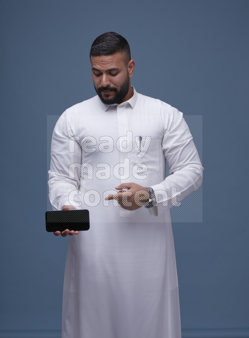 A man Showing his phone in Blue Background wearing Saudi Thob