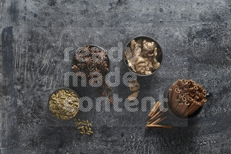 Ginger, Cardamom, Star anise and cinnamon sticks in 4 bowls on a textured black background