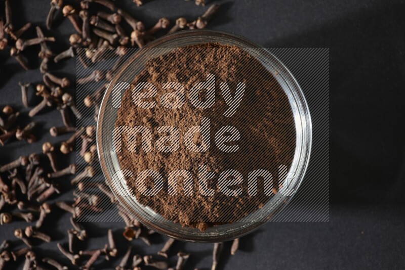 وعاء زجاجي ممتلئ ببودرة القرنفل مع حبوب القرنفل المتناثرة على أرضية سوداء