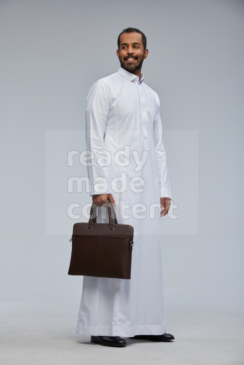 Saudi man wearing thob standing holding suitcase on gray background