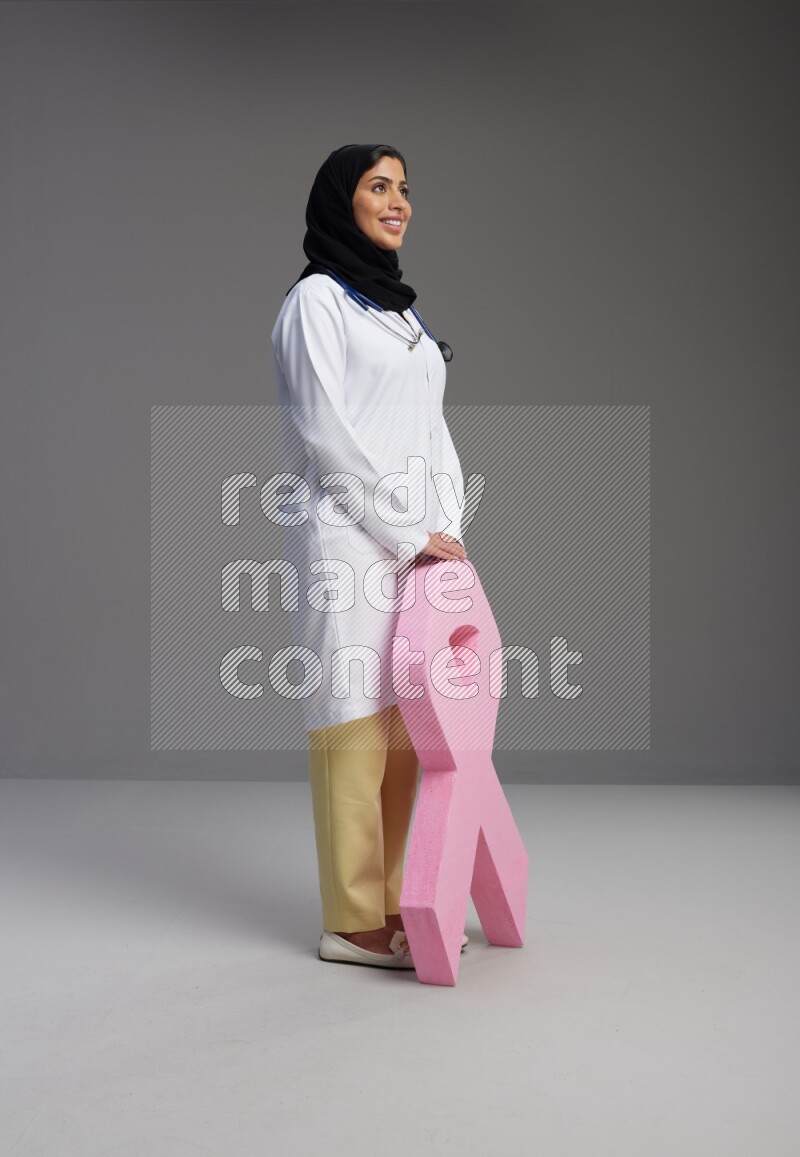 Saudi woman wearing lab coat with stethoscope standing holding awareness ribbon symbol standing on Gray background