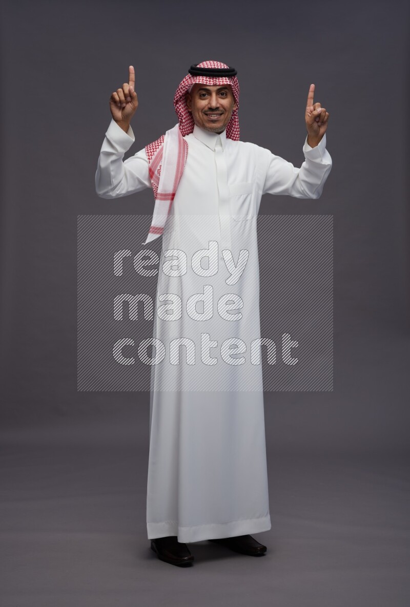 Saudi man wearing thob and shomag standing interacting with the camera on gray background