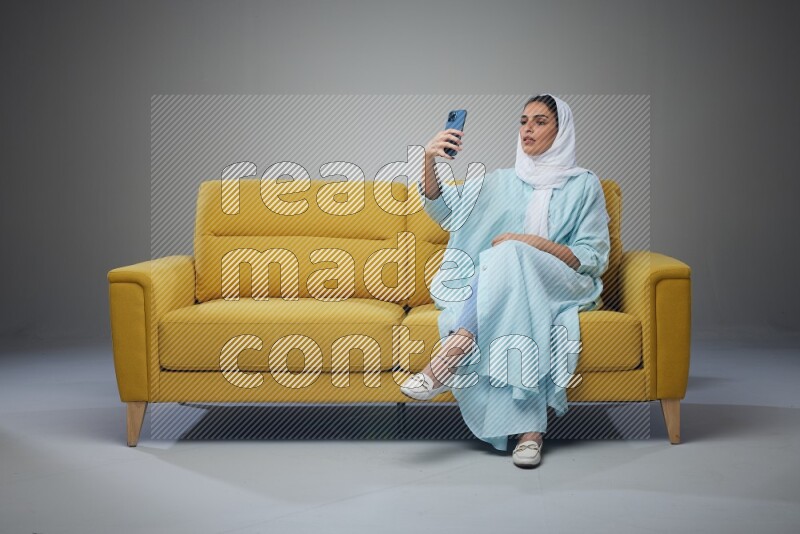 A Saudi woman wearing a light blue Abaya and white head scarf sitting on a yellow sofa and taking selfies with her phone eye level on a grey background