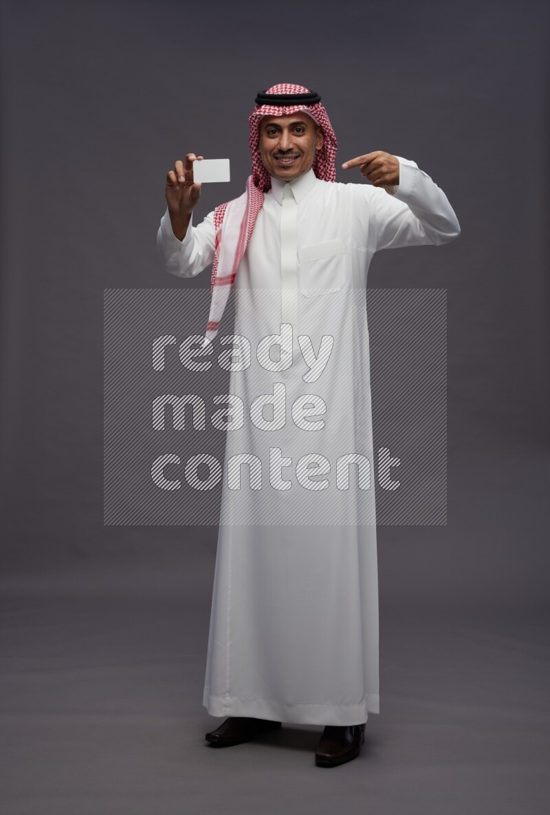 Saudi man wearing thob and shomag standing holding ATM card on gray background