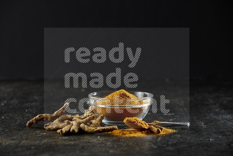 وعاء زجاجي وملعقة معدنية ممتلئان ببودرة الكركم وأصابع الكركم المجففة بجانبهما على خلفية سوداء