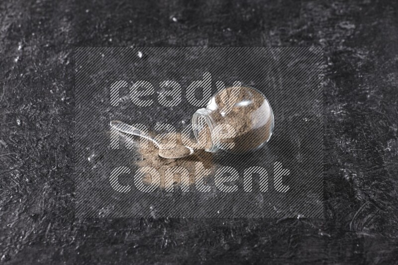 A flipped glass spice jar full of black pepper powder with a metal spoon full of powder on textured black flooring