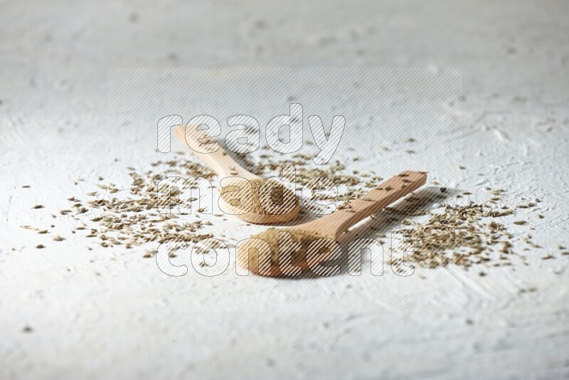 2 Wooden spoons full of cumin powder and cumin seeds on textured white flooring
