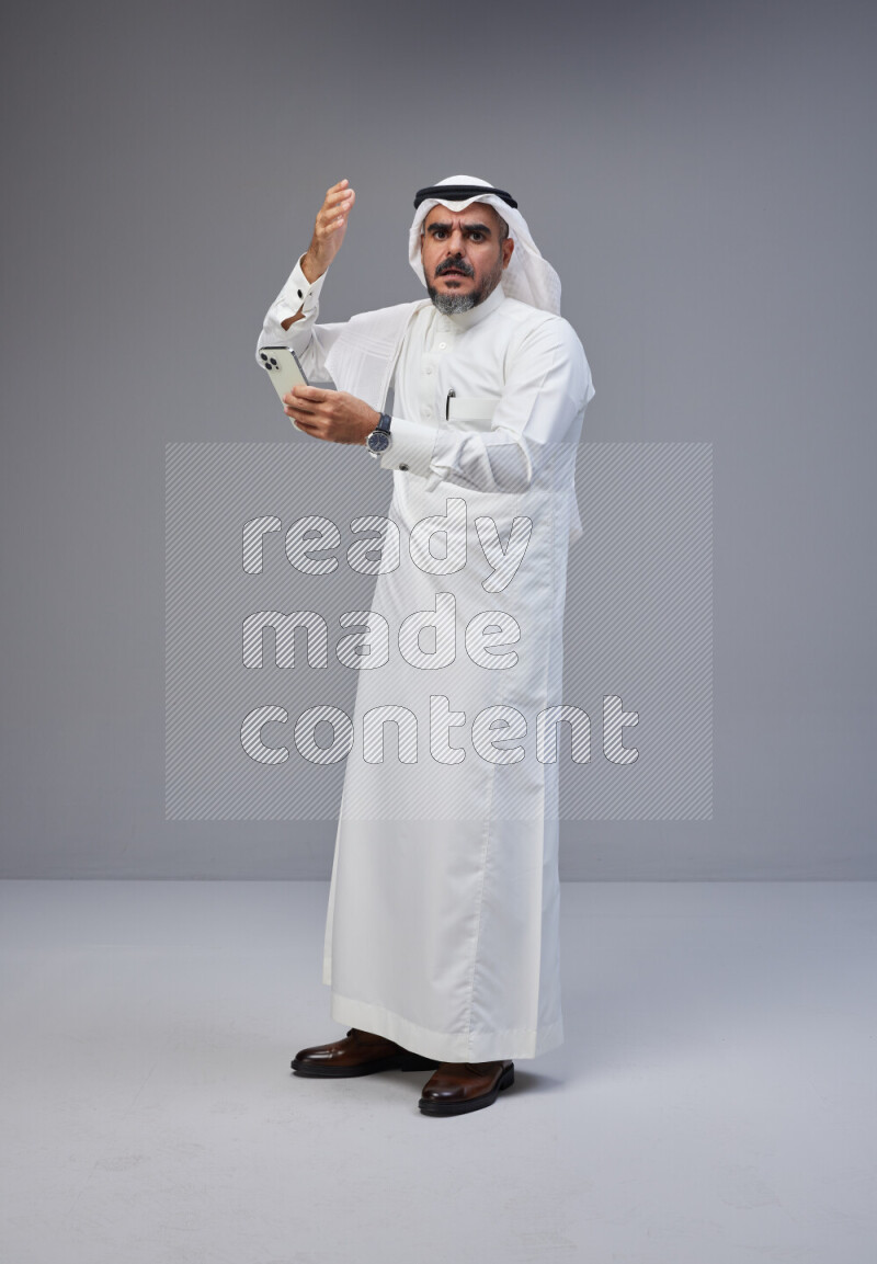 Saudi man Wearing Thob and white Shomag standing texting on phone on Gray background