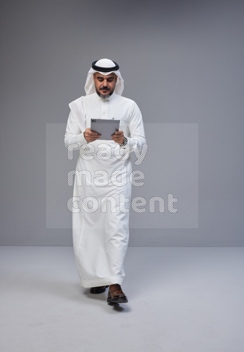Saudi man Wearing Thob and white Shomag standing working on tablet on Gray background