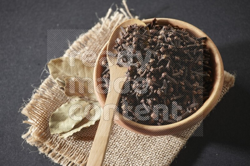 A wooden bowl, a wooden spoon full of cloves, and bay leaves (laurel) on a piece of burlap on a black flooring