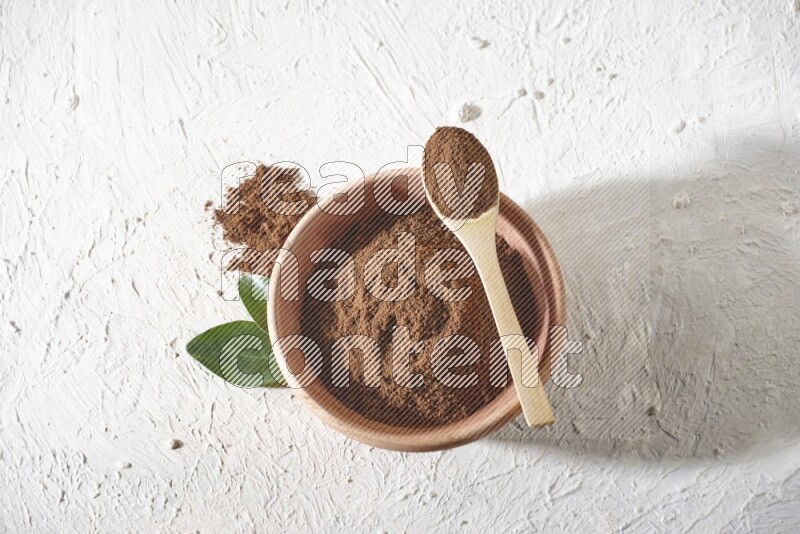 A wooden bowl and a wooden spoon full of cloves powder on a textured white flooring