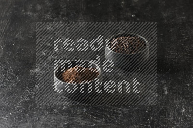 2 Black pottery bowls full of cloves and the other full of cloves powder on textured black flooring
