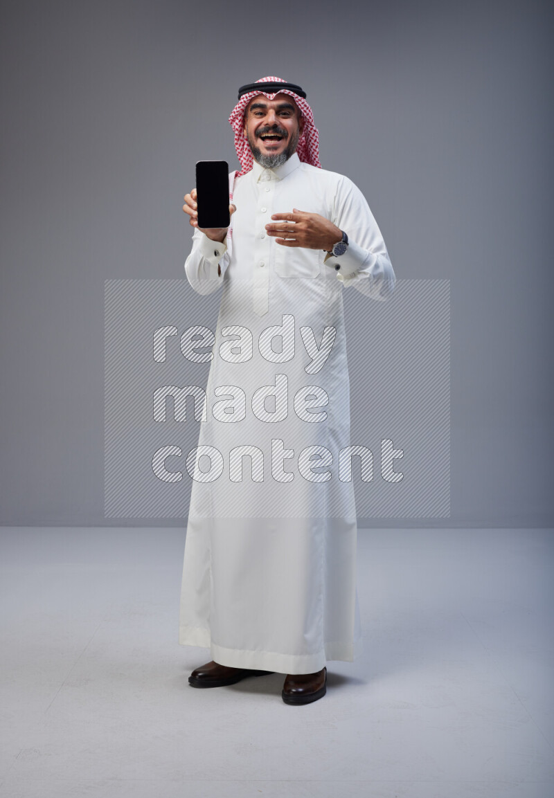 Saudi man Wearing Thob and red Shomag standing showing phone to camera on Gray background