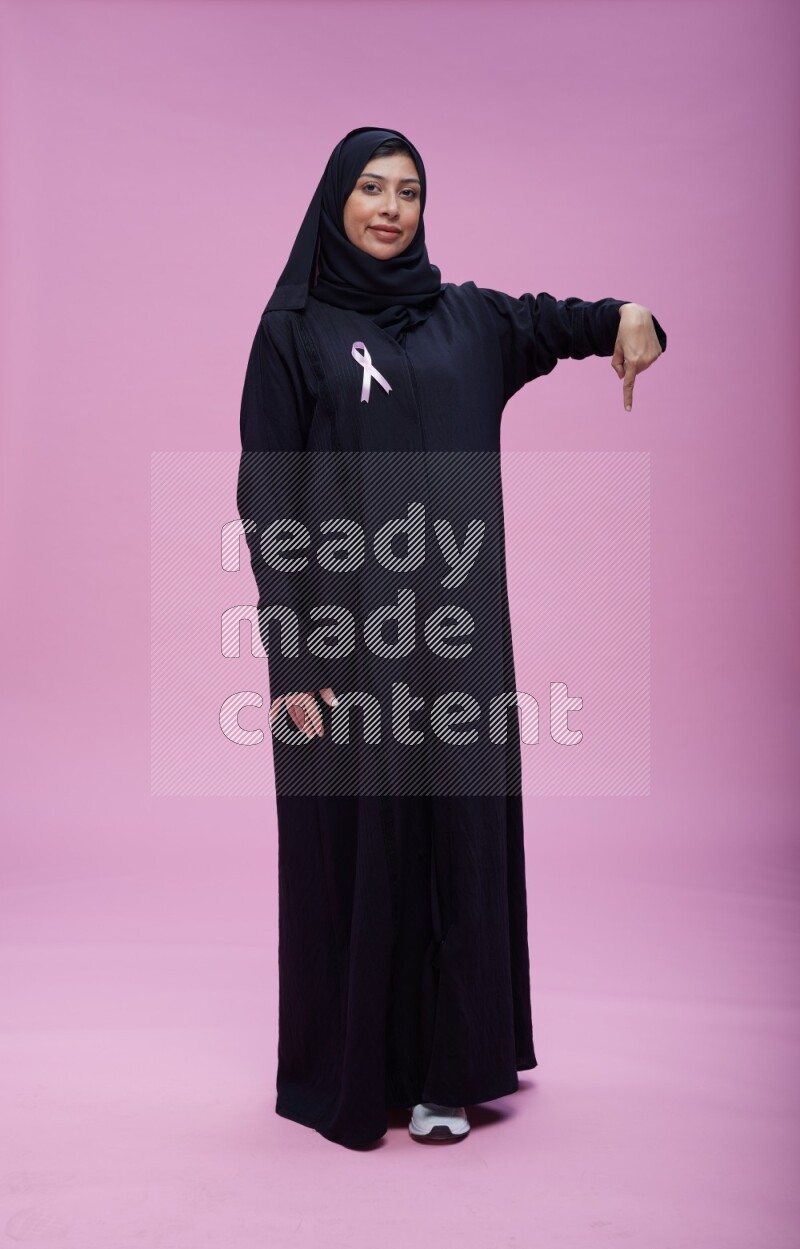 Saudi woman wearing pink ribbon on Abaya standing interacting with the camera on pink background