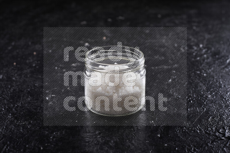 A glass jar full of coarse sea salt crystals on black background