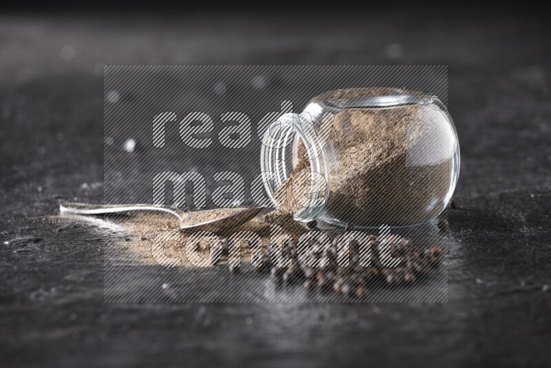 A flipped glass spice jar full of black pepper powder with a metal spoon full of powder and pepper beads on textured black flooring